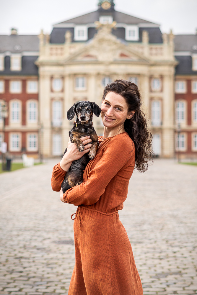Foto von Lisa Werner Fotografie, Hund und Mensch, Frau steht vor einem Schloss und hält Dackel im Arm