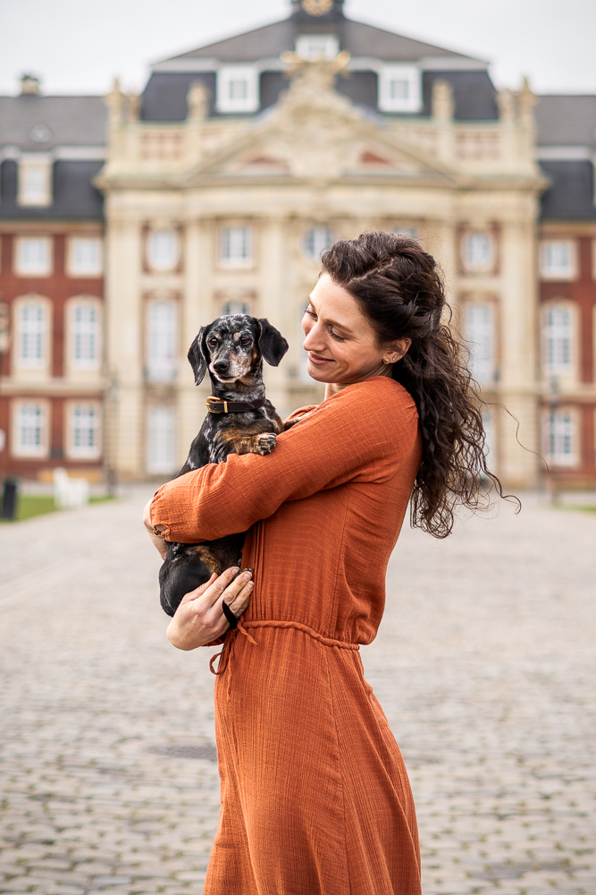 Foto von Lisa Werner Fotografie, Hund und Mensch, Frau steht vor einem Schloss und hält Dackel im Arm