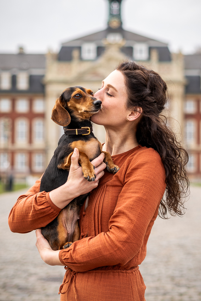 Foto von Lisa Werner Fotografie, Hund und Mensch, Frau steht vor einem Schloss und hält Dackel im Arm