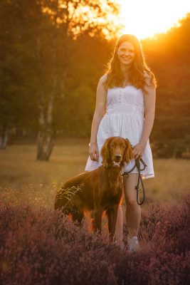 Foto von Lisa Werner Fotografie, Hund und Mensch, Frau und Irish Red Setter stehen in lila Heideblüten im Sonnenuntergang